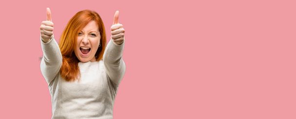Beautiful young redhead woman stand happy and positive with thumbs up approving with a big smile expressing okay gesture - Фото, зображення