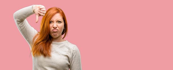 Kaunis nuori punapää nainen osoittaa peukalot alas onneton merkki inhoa, negatiivinen ilme ja paheksuntaa
 - Valokuva, kuva