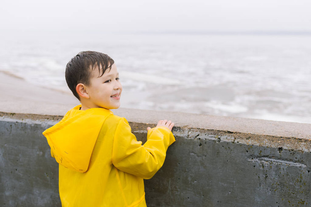 Ένα αγόρι σε ένα κίτρινο αδιάβροχο φαίνεται στην επιφάνεια του νερού (λίμνη, θάλασσα, ποτάμι, ωκεανός). Νεφελώδης καιρός, βροχή, εκτός σπιτιού, με τα πόδια. - Φωτογραφία, εικόνα