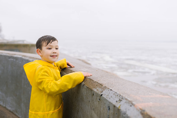 A boy in a yellow raincoat looks at the water surface (lake, sea, river, ocean). Overcast weather, rain, outdoor, walk. - Foto, imagen
