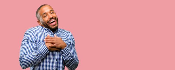 Hombre afroamericano con barba que tiene una sonrisa encantadora cogido de la mano en el corazón con ganas de mostrar amor y simpatía
 - Foto, imagen