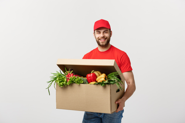 Concepto de entrega: Hermoso hombre de mensajero de entrega de comestibles caucásicos en uniforme rojo con caja de comestibles con frutas y verduras frescas
 - Foto, imagen