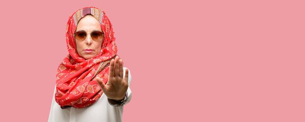 Mujer árabe musulmana de mediana edad con hiyab molesto con mala actitud haciendo señal de stop con la mano, diciendo que no, expresando seguridad, defensa o restricción, tal vez empujando
 - Foto, imagen