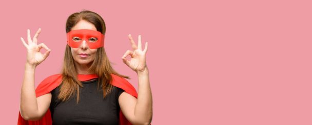 Жінка-супергерой середнього віку в червоній масці та накидці робить жест зі знаком ок з обома руками, виражаючи медитацію та релаксацію
 - Фото, зображення