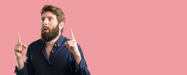 Young hipster man with big beard happy and surprised cheering expressing wow gesture pointing up - Photo, image