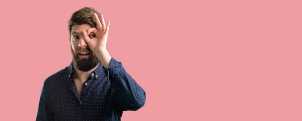 Mladí bokovky muž s bradáči při pohledu na fotoaparát skrze prsty v pořádku gestu. Napodobovat dalekohled, krásné oči a úsměv - Fotografie, Obrázek