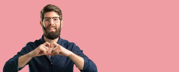 junger Hipster-Mann mit dickem Bart zeigt glücklich Liebe mit Händen in Herzform, die ein gesundes und eheliches Symbol ausdrücken - Foto, Bild