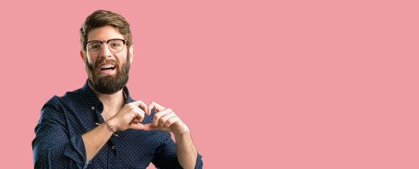 Jeune homme hipster avec une grande barbe heureux montrant l'amour avec les mains en forme de coeur exprimant symbole de santé et de mariage
 - Photo, image