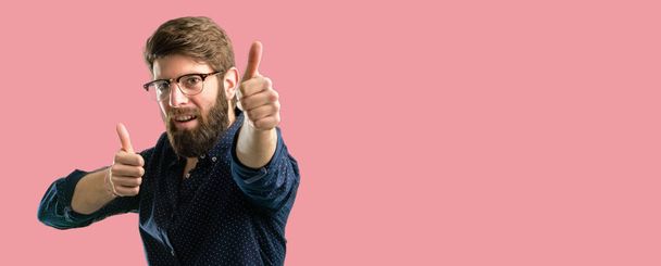 Homem jovem hipster com barba grande ficar feliz e positivo com polegares para cima aprovar com um grande sorriso expressando gesto ok
 - Foto, Imagem