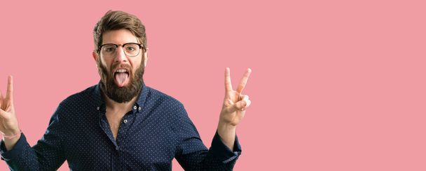 Young hipster man with big beard looking at camera showing tong and making victory sign with fingers - Photo, Image