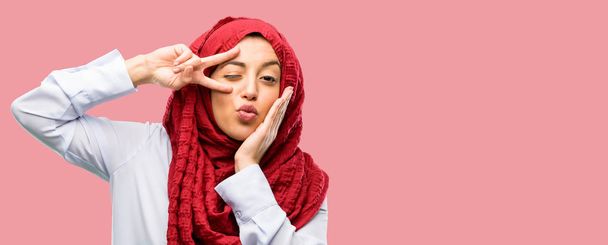 Młoda kobieta Arabska noszenie hidżabu patrząc na kamery przez jej palce w geście zwycięstwa mrugając okiem i Buziak - Zdjęcie, obraz
