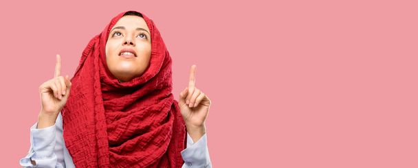 junge arabische Frau im Hijab glücklich und überrascht jubelnd nach oben zeigend - Foto, Bild