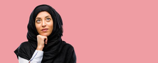 Mujer árabe joven usando hijab pensando y mirando hacia arriba expresando duda y asombro
 - Foto, Imagen