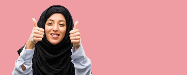 Jonge Arabische vrouw het dragen van hijab glimlachend in grote lijnen weergegeven: duimschroef opwaarts gebaar camera, expressie van en goedkeuring - Foto, afbeelding