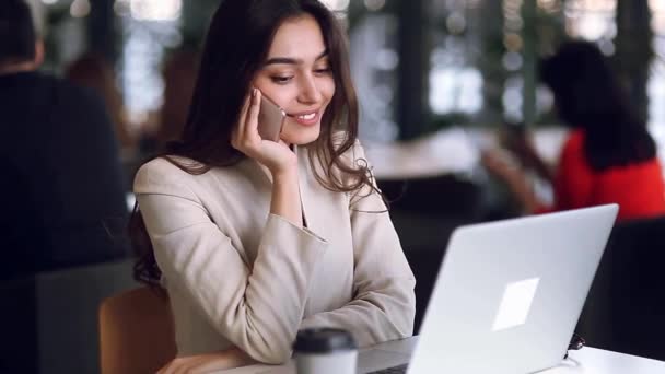 Beautiful business girl working on the phone at work on a laptop, enjoying the success. Conclusion of the contract, communication online. The modern woman is a freelancer working in a cafe - Metraje, vídeo