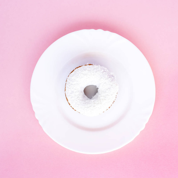 Donut covered with white glaze and sprinkled with coconut shavings on white plate on pink background - Foto, Bild