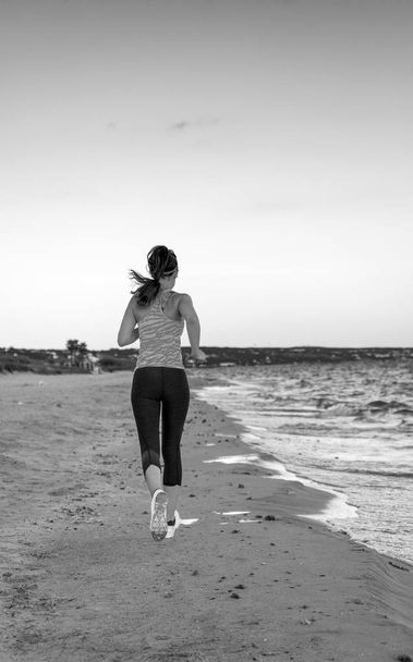 Visto desde atrás, una mujer sana trotando en ropa deportiva en la orilla del mar por la noche corriendo.
 - Foto, Imagen