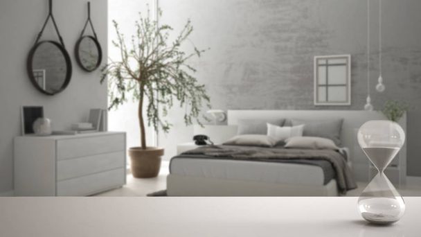 Mesa blanca o estante con reloj de arena de cristal que mide el paso del tiempo sobre un dormitorio moderno borroso con olivo, diseño de interiores de arquitectura, espacio de copia de fondo
 - Foto, imagen