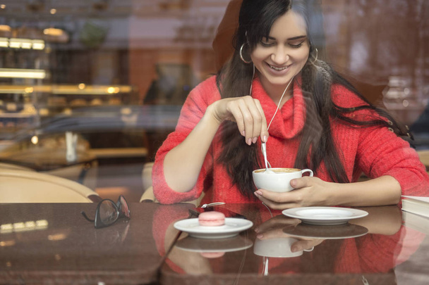 Cute brunette plus size woman working on a distance (freelance), sitting in cafe, drinking coffee and eating macaron. She wearing red sweater, listen music and typing messages on her phone - Valokuva, kuva