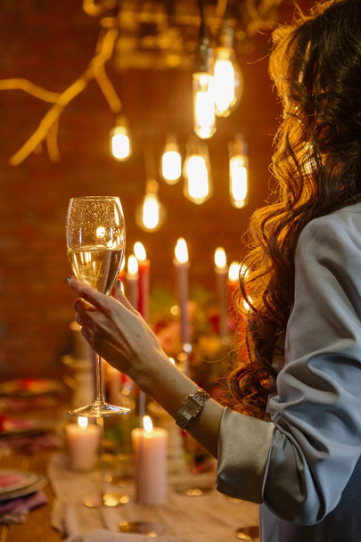 Woman standing neat wedding table, celebrate with a glass of wine in her hand. No face. Decor, candles and edison lamps on a background - Фото, изображение