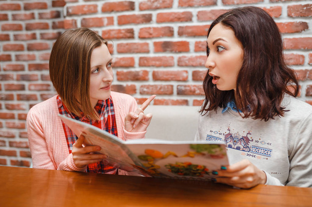 Dos chicas amigas en un café leyendo el menú con emociones divertidas
 - Foto, Imagen