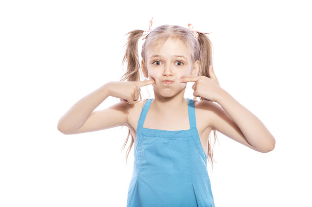 Young seven years old brunette girl in blue dress on a white isolated background. She blows her cheeks, funny emotions on her face - Photo, Image