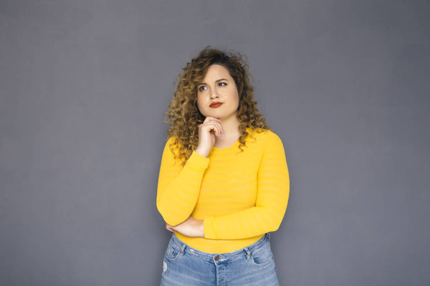 Cute brunette plus size woman with curly hair in yellow sweater and jeans standing on a neutral grey background. She was thinking, thoughtful emotion on her face. Copy space - Foto, afbeelding