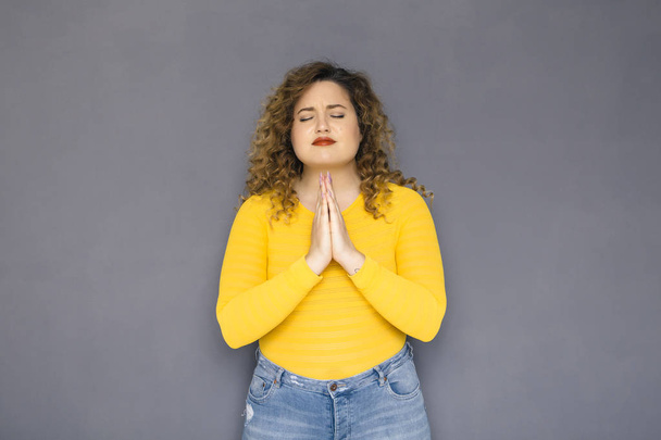 Cute brunette plus size woman with curly hair in yellow sweater and jeans standing on a neutral grey background. She makes a prayer gesture or sign with her hands - Zdjęcie, obraz