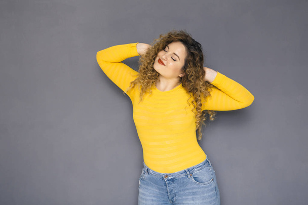 Cute brunette plus size woman with curly hair in yellow sweater and jeans standing on a neutral grey background. She stretched herself - Photo, Image