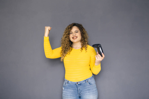 Cute brunette plus size woman with curly hair in yellow sweater and jeans standing on a neutral grey background. She holds a joystick, playing on console games - Photo, Image