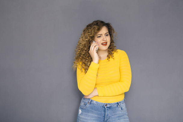Χαριτωμένο μελαχρινή συν μέγεθος γυναίκα με σγουρά μαλλιά σε κίτρινο πουλόβερ και τζιν που στέκεται πάνω σε ένα ουδέτερο γκρι παρασκήνιο. Μιλά στο κινητό της τηλέφωνο, τηλεφωνικές κλήσεις. Χώρο αντίγραφο - Φωτογραφία, εικόνα