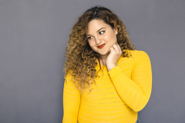 Cute brunette plus size woman with curly hair in yellow sweater and jeans standing on a neutral grey background. She smiles happily looking sideways - Valokuva, kuva