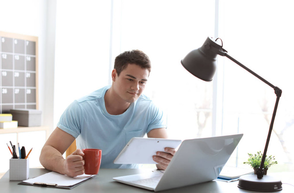 Портрет уверенного молодого человека с планшетом и ноутбуком и чашкой за столом
 - Фото, изображение