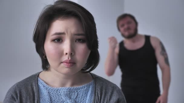 Retrato de una triste chica asiática deprimida, marido borracho en el fondo está de duelo, alcohol, violencia, mira a la cámara. 60 fps
 - Metraje, vídeo