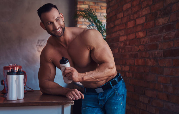 Portrait of a Tall stylish shirtless bodybuilder dressed in jeans, prepares protein mixture before morning exercise, standing in a kitchen with a loft interior, looking out the window. - Photo, Image