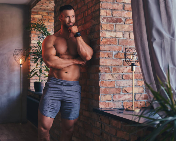 Grand bodybuilder torse nu élégant habillé en short de sport, debout dans une chambre avec un intérieur loft, appuyé contre un mur de briques, regardant par la fenêtre
. - Photo, image