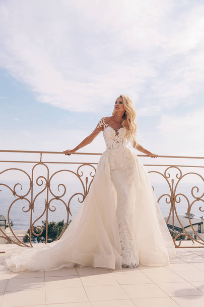 fashion photo of beautiful sensual woman with blond hair in luxurious wedding dress and accessories posing outdoor with sea and mountains view - Fotó, kép