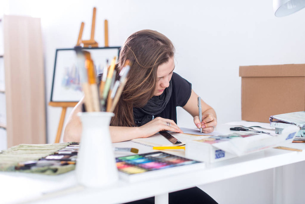 Νεαρή γυναίκα καλλιτέχνης σχέδιο σκίτσο χρησιμοποιώντας sketchbook με μολύβι στο χώρο εργασίας της στο στούντιο. Πλευρά άποψη πορτρέτο του εμπνευσμένος ζωγράφος. - Φωτογραφία, εικόνα