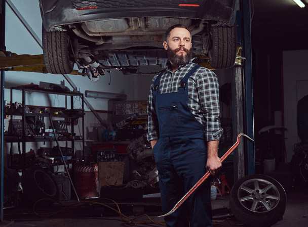 mecánico experto barbudo vestido con un uniforme, de pie con una palanca en la mano, contra un coche en un ascensor en el garaje
. - Foto, Imagen