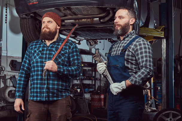 zwei bärtige brutale Mechaniker reparieren ein Auto auf einem Aufzug in einer Garage. - Foto, Bild