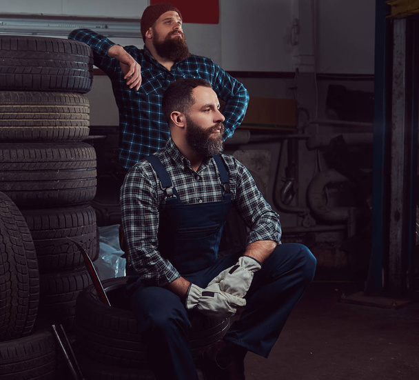 Due meccanici barbuti vicino a una pila di vecchie gomme da auto in garage
. - Foto, immagini