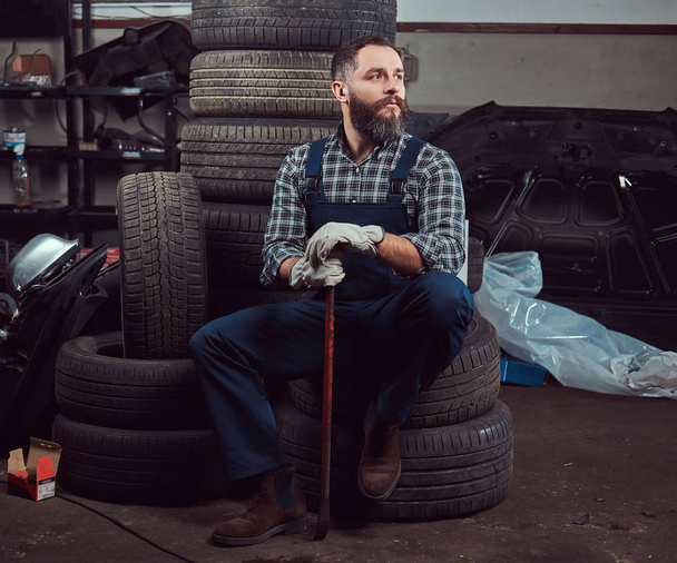 Mécanicien barbu vêtu d'un uniforme, tient un pied-de-biche, s'assoit sur de vieux pneus de voiture dans le garage
. - Photo, image