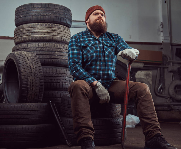 Brodaty mechanik brutalny, ubrany w koszulę Polar i czapka, posiada Łom, siada na zużytych opon samochodowych w garażu. - Zdjęcie, obraz