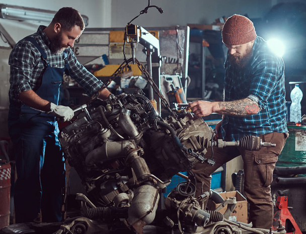 Два бородатых специалиста-механика ремонтируют двигатель автомобиля, который поднимается на гидравлическом подъемнике в гараже. Сервисная станция
. - Фото, изображение