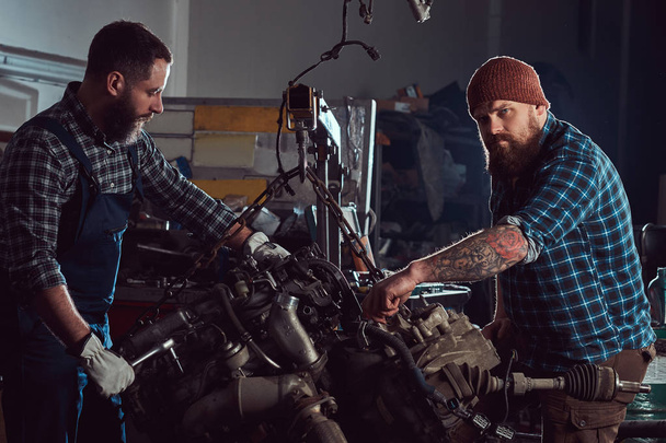 Kaksi parrakas mekaniikka asiantuntija korjaa auton moottori, joka nostetaan hydraulinen hissi autotallissa. Huoltoasema
. - Valokuva, kuva