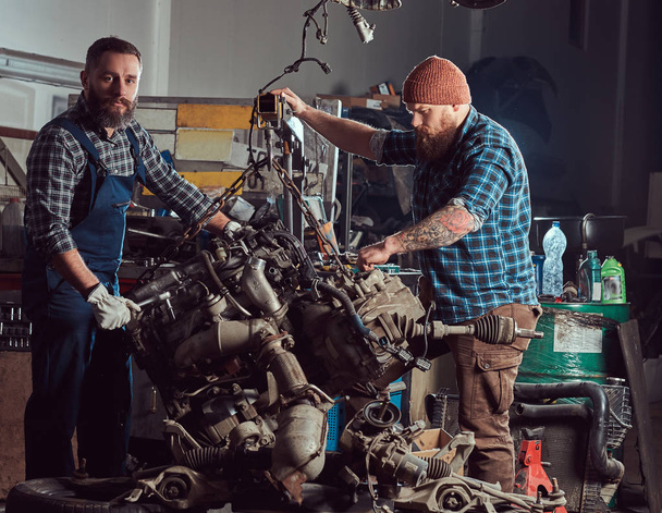 Два бородатих механіка фахівець ремонтує автомобільний двигун, який піднімається на гідравлічний підйомник в гаражі. Станція обслуговування
. - Фото, зображення