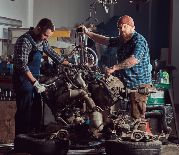 Two bearded mechanics specialist repairs the car engine which is raised on the hydraulic lift in the garage. Service station. - Fotó, kép