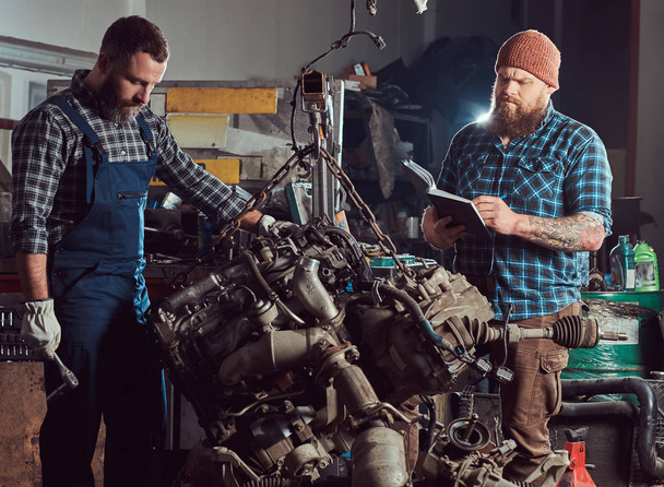 Bearded expert mechanic records results of inspecting a car while his partner checking a problem with a car engine. A scene from a service station. - Photo, Image