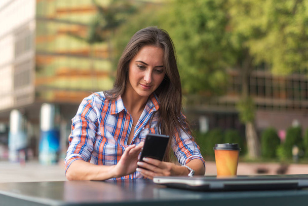 Viehättävä nuori nainen, joka lukee tekstiviestiä kännykällään. Tyttö istuu ulkona älypuhelimella
 - Valokuva, kuva