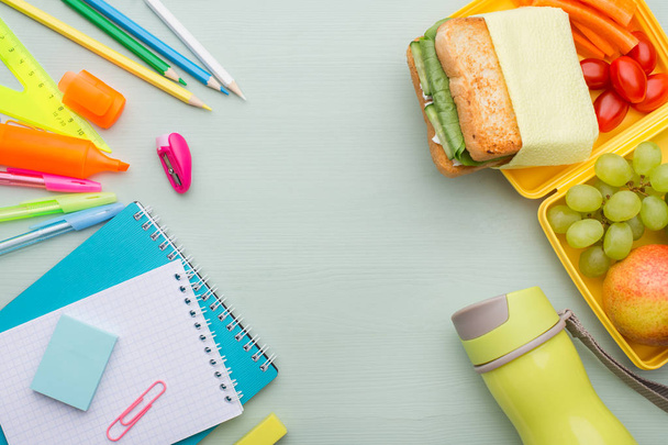 A pile of various stationery on a wooden blue table, a notepad, colored pencils, ruler, marker, planer, space for text. Delicious school lunch box, sandwich and fruit. The concept of preparing children for school. - Photo, image